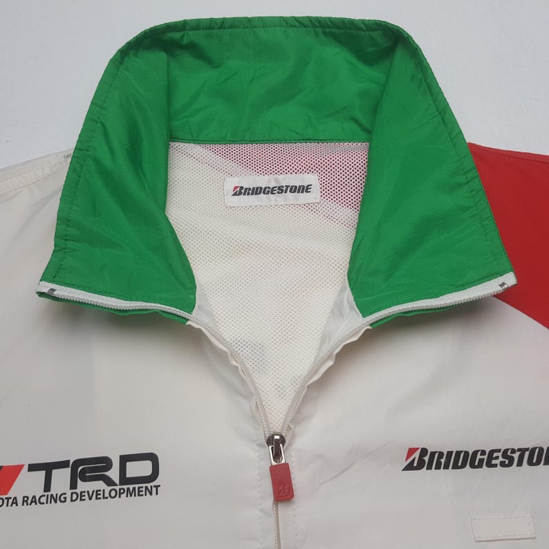Puede incluir: Chaqueta de carreras blanca y roja con cuello verde. La chaqueta presenta los logotipos "TRD TOYOTA RACING DEVELOPMENT" y "BRIDGESTONE" en negro. La chaqueta tiene una cremallera completa y un forro de malla blanca.