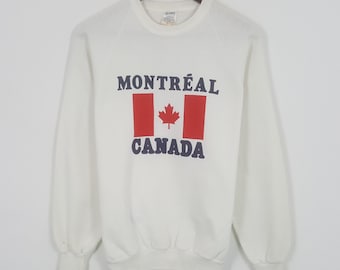 May include: White sweatshirt with long sleeves. The front features the text "MONTREAL CANADA" in navy blue, with a red and white Canadian flag design in the center. The sweatshirt is hanging on a wooden hanger.