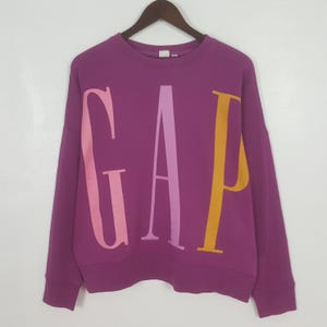 Peut inclure: Un sweat-shirt violet avec le mot "GAP" en grandes lettres colorées sur le devant. Les lettres sont dans des tons de rose et de jaune. Le sweat-shirt est suspendu à un cintre en bois.