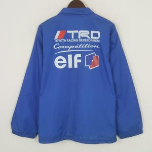 May include: A blue jacket with the text "TRD TOYOTA RACING DEVELOPMENT Compétition elf" printed on the back in white, red, and blue. The jacket has a collared neckline and long sleeves.