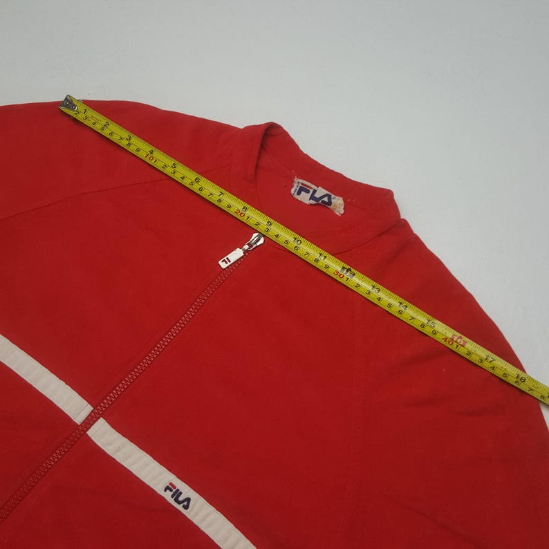 May include: Red zip-up jacket with white stripes and the "FILA" logo. The jacket has a white zipper and a small "FILA" logo on the collar. A yellow measuring tape is laid across the jacket.