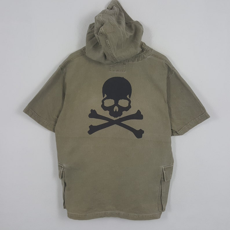 Puede incluir: Camiseta con capucha de manga corta verde oliva con un gr&aacute;fico de calavera y huesos cruzados negros en la espalda. La camiseta tiene dos bolsillos delanteros y una capucha. La camiseta est&aacute; hecha de tela duradera.