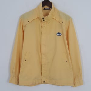 May include: A light yellow jacket with a high collar and long sleeves. The jacket has two front pockets and a small Audi logo on the chest. The jacket is buttoned up the front and has a simple, classic design.