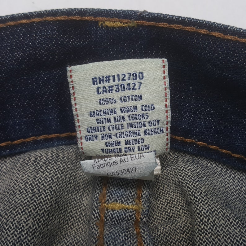 May include: Close-up of a denim garment tag with text. The tag reads "RN#112790 CA#30427 100% COTTON MACHINE WASH COLD WITH LIKE COLORS GENTLE CYCLE INSIDE OUT ONLY NON-CHLORINE BLEACH WHEN NEEDED TUMBLE DRY LOW MADE IN EUA Fabrique AU EUA CA#30427".
