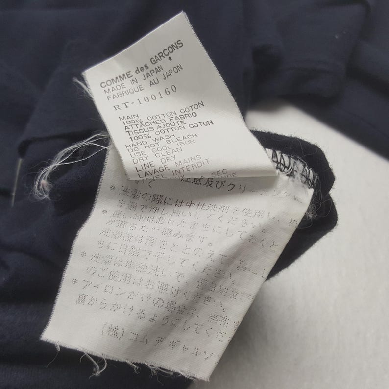 K&ouml;nnte beinhalten: Nahaufnahme eines marineblauen Kleidungsst&uuml;cketiketts mit wei&szlig;em Text. Das Etikett lautet "COMME des GARCONS MADE IN JAPAN FABRIQUE AU JAPON" und enth&auml;lt Pflegehinweise in Englisch und Japanisch. Das Kleidungsst&uuml;ck besteht aus 100 % Baumwolle.