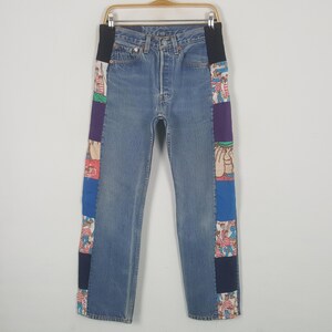 Könnte beinhalten: Hellblaue Jeans mit Patchwork-Einsätzen an den Seiten. Der Patchwork-Stoff weist verschiedene Farben und Muster auf, darunter Cartoonfiguren. Die Jeans hängen an einem Holzbügel.