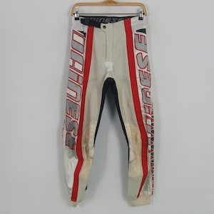 Peut inclure: Pantalon de moto cr&egrave;me avec des d&eacute;tails rouges et blancs. Le pantalon porte le mot "DAINESE" en grandes lettres rouges sur le c&ocirc;t&eacute; et un panneau noir &agrave; l'int&eacute;rieur de la jambe. Suspendu &agrave; un cintre en bois.