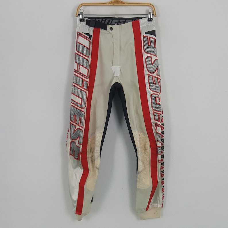 Peut inclure: Pantalon de moto cr&egrave;me avec des d&eacute;tails rouges et blancs. Le pantalon porte le mot "DAINESE" en grandes lettres rouges sur le c&ocirc;t&eacute; et un panneau noir &agrave; l'int&eacute;rieur de la jambe. Suspendu &agrave; un cintre en bois.