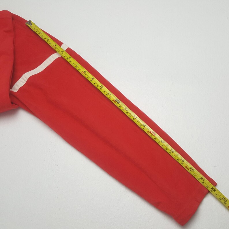 May include: A red long-sleeved garment with a white stripe detail. A yellow and black measuring tape is laid across the garment, showing measurements in inches.