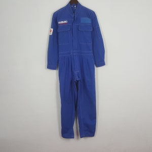 Może przedstawiać: Niebieski kombinezon z białymi przeszyciami, z logo "SUZUKI" na piersi i rękawie. Jednoczęściowy strój ma wiele kieszeni i zapięcie na zamek błyskawiczny, zawieszony na gładkim tle.
