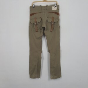 May include: Khaki-colored pants with brown leather accents and multiple pockets. The pants have a straight leg design and are hanging on a metal hanger. The back of the pants features two large pockets with leather straps.