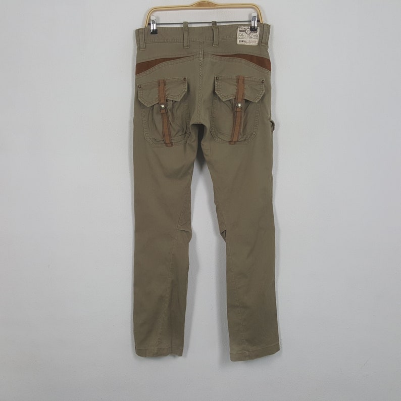 May include: Khaki-colored pants with brown leather accents and multiple pockets. The pants have a straight leg design and are hanging on a metal hanger. The back of the pants features two large pockets with leather straps.
