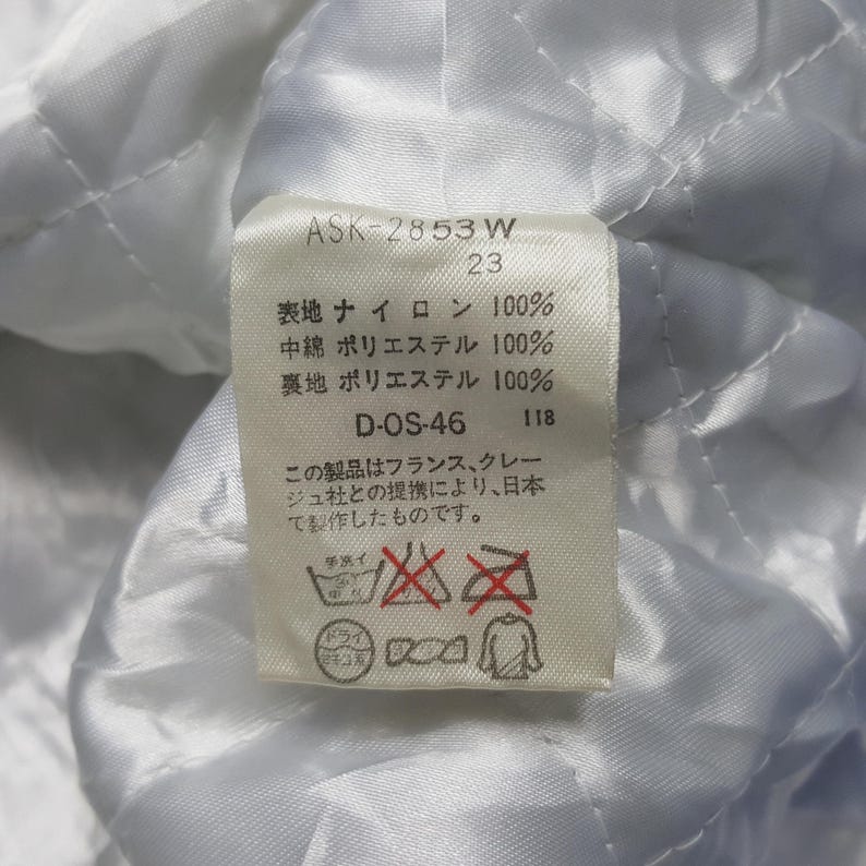 May include: Close-up of a white fabric tag with Japanese text and care symbols. The tag includes the text "ASK-2853 W" and "D-OS-46". The fabric appears to be quilted.