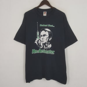 May include: Black t-shirt featuring a graphic from the movie "Re-Animator." The design includes an illustration of a man with glasses holding a syringe, with the text "Herbert West..." above and "Re-Animator" below in green.