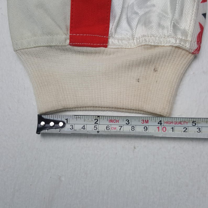 Peut inclure: Gros plan sur un poignet de manche blanc et rouge avec un poignet en tricot c&ocirc;tel&eacute; cr&egrave;me. La manchette est mesur&eacute;e avec un m&egrave;tre ruban, environ 5 cm de large.