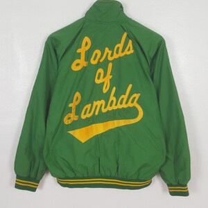 Może przedstawiać: Zielona kurtka bomberka z żółtym napisem "Lords of Lambda" na plecach. Kurtka ma prążkowany kołnierz, mankiety i dół z żółto-zielonymi paskami. Wykonana z lekkiego materiału.
