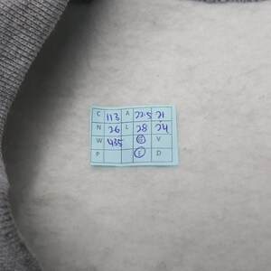 Pode incluir: Uma foto em close-up de uma pe&ccedil;a de roupa cinza com interior cinza claro. Uma pequena etiqueta azul retangular est&aacute; afixada &agrave; pe&ccedil;a de roupa, exibindo medidas e letras manuscritas. O texto da etiqueta &eacute; parcialmente vis&iacute;vel.