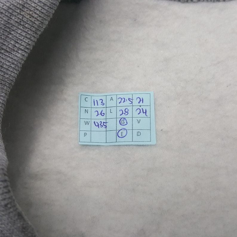 Pode incluir: Uma foto em close-up de uma pe&ccedil;a de roupa cinza com interior cinza claro. Uma pequena etiqueta azul retangular est&aacute; afixada &agrave; pe&ccedil;a de roupa, exibindo medidas e letras manuscritas. O texto da etiqueta &eacute; parcialmente vis&iacute;vel.