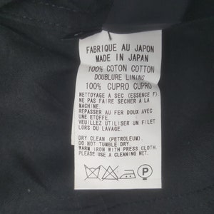 May include: A white tag with black text, including "FABRIQUE AU JAPON" and "MADE IN JAPAN". The tag details fabric composition, care instructions, and cleaning symbols. The tag is against a dark background.