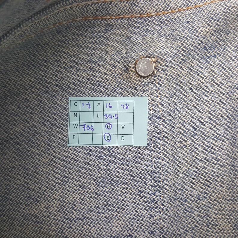 May include: Close-up of a textured denim fabric in shades of blue and grey. A small, silver button is visible near the top. A rectangular tag with handwritten numbers and letters is attached to the fabric.