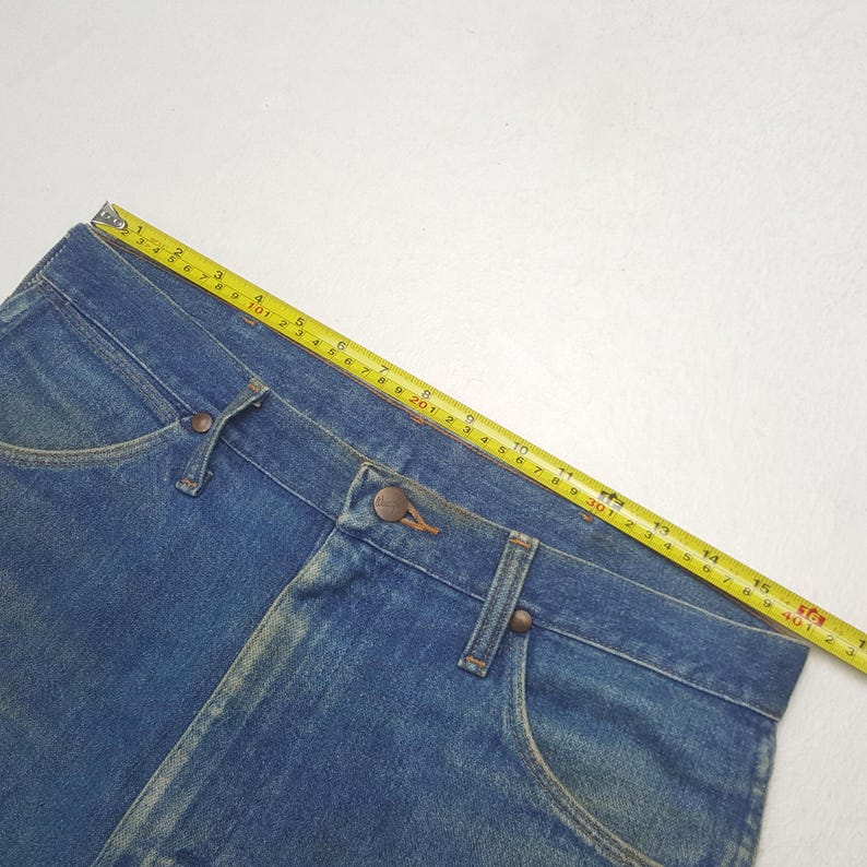 May include: Close-up of blue denim jeans with a yellow measuring tape laid across the waistband. The jeans have a button closure and classic pocket design. The measuring tape shows measurements in centimetres.