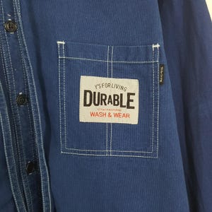 May include: Close-up of a navy blue button-down shirt. The shirt features a white stitched pocket with the text "Y'S FOR LIVING DURABLE WASH & WEAR". The shirt has black buttons and white stitching details.