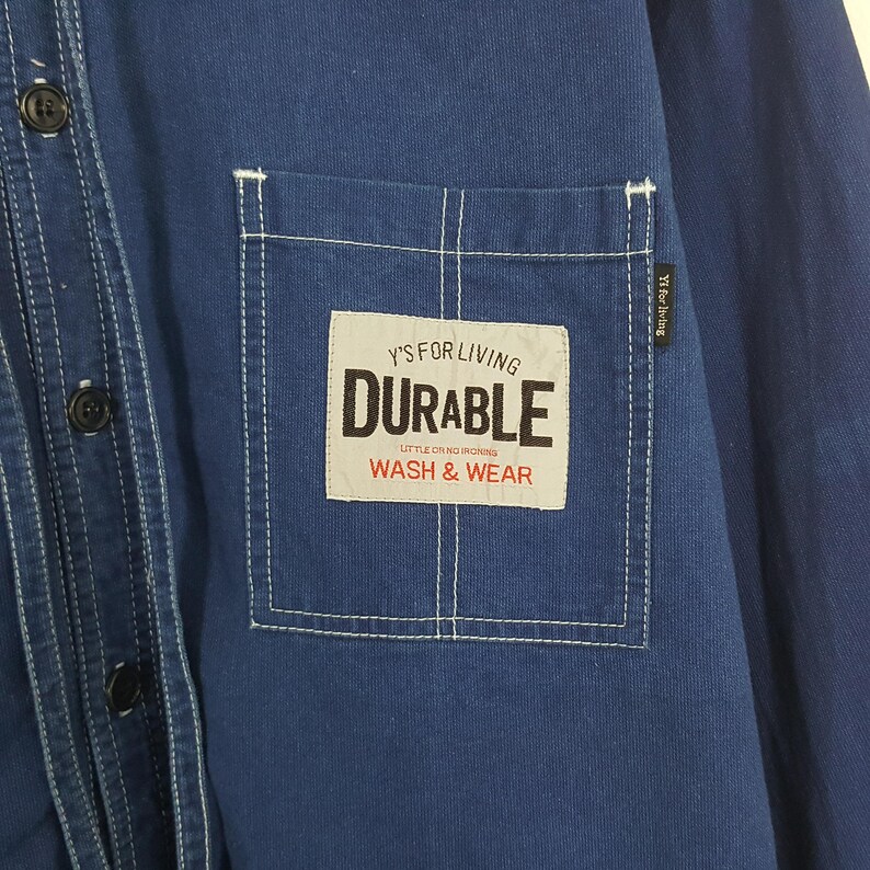 May include: Close-up of a navy blue button-down shirt. The shirt features a white stitched pocket with the text "Y'S FOR LIVING DURABLE WASH & WEAR". The shirt has black buttons and white stitching details.