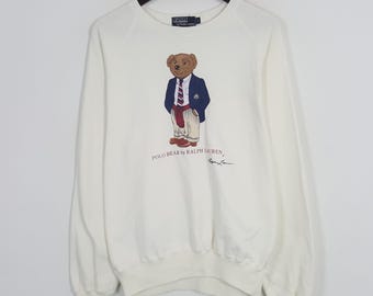 Puede incluir: Sudadera de color crema con un gráfico de Polo Bear. El oso viste una blazer azul marino, corbata a rayas y pantalones rojos. El texto "POLO BEAR by RALPH LAUREN" está impreso debajo. La sudadera está colgada en una percha negra.