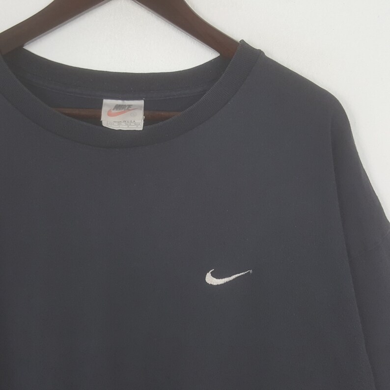 Peut inclure: T-shirt noir &agrave; col rond avec un petit logo Nike blanc sur la poitrine gauche. Le t-shirt est suspendu &agrave; un cintre en bois sur fond blanc. Le logo Nike est brod&eacute;.