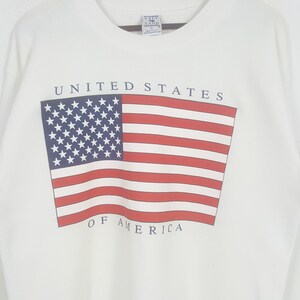 May include: White sweatshirt featuring the American flag design. The flag has red and white stripes with a blue square containing white stars. The words "UNITED STATES" and "OF AMERICA" are printed above and below the flag.