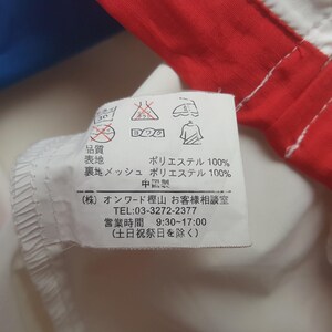 Puede incluir: Primer plano de una etiqueta de tela blanca con texto japon&eacute;s y s&iacute;mbolos de cuidado. La etiqueta est&aacute; adherida a una prenda roja y blanca. El texto incluye la composici&oacute;n de la tela e informaci&oacute;n de la empresa.