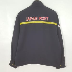 Puede incluir: Chaqueta azul marino con mangas largas. La parte trasera de la chaqueta presenta el texto "JAPAN POST" en letras rojas sobre una franja amarilla. Un pequeño parche de la bandera japonesa está en la manga derecha.