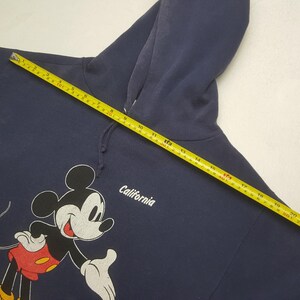 Puede incluir: Sudadera con capucha azul marino con un gr&aacute;fico cl&aacute;sico de Mickey Mouse en negro, blanco y rojo. La palabra "California" est&aacute; impresa en blanco. La sudadera tiene un cord&oacute;n y una capucha.