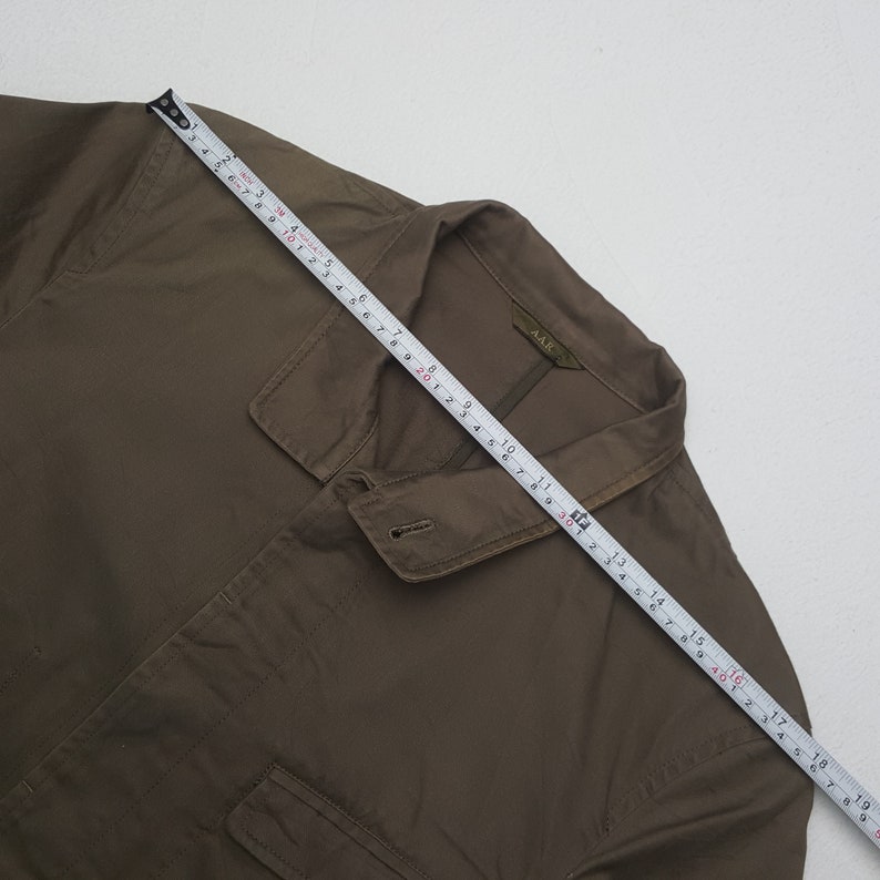 Puede incluir: Camisa verde oliva con cuello, cierre de botones y dos bolsillos delanteros. Una cinta m&eacute;trica est&aacute; colocada sobre la camisa, mostrando medidas en cent&iacute;metros. La camisa tiene el texto "AAR" en el cuello.