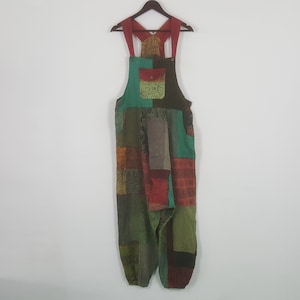 May include: Patchwork overalls in shades of green, red, brown, and orange hang on a wooden hanger. The overalls feature a bib pocket and adjustable straps. The fabric is a mix of patterned and solid patches.