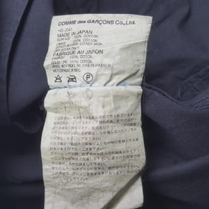 Puede incluir: Una etiqueta de Comme des Gar&ccedil;ons Co., Ltd. con texto en ingl&eacute;s y japon&eacute;s. La etiqueta indica que el art&iacute;culo est&aacute; hecho en Jap&oacute;n con 100% algod&oacute;n. Incluye instrucciones de cuidado, como limpieza en seco y no planchar.