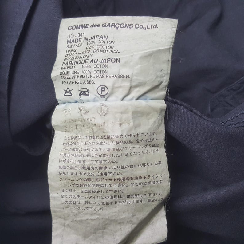 Puede incluir: Una etiqueta de Comme des Gar&ccedil;ons Co., Ltd. con texto en ingl&eacute;s y japon&eacute;s. La etiqueta indica que el art&iacute;culo est&aacute; hecho en Jap&oacute;n con 100% algod&oacute;n. Incluye instrucciones de cuidado, como limpieza en seco y no planchar.