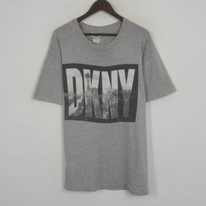 May include: A grey DKNY t-shirt featuring a black and white graphic of the New York City skyline and the Statue of Liberty. The word "DKNY" is printed in large, bold, white letters over the image. The shirt has short sleeves and a crew neck.