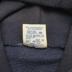 Puede incluir: Primer plano de una sudadera con capucha azul marino con una etiqueta blanca. La etiqueta muestra las instrucciones de lavado y la composici&oacute;n de la tela: 50% algod&oacute;n y 50% poli&eacute;ster. Tambi&eacute;n se ve el logotipo y la direcci&oacute;n de Russell Corp.