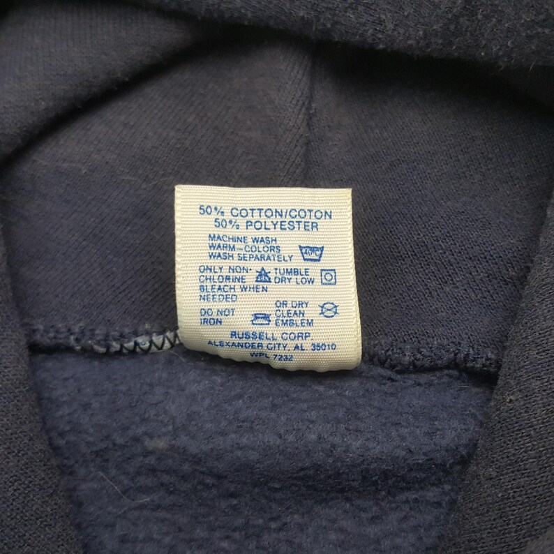 Puede incluir: Primer plano de una sudadera con capucha azul marino con una etiqueta blanca. La etiqueta muestra las instrucciones de lavado y la composici&oacute;n de la tela: 50% algod&oacute;n y 50% poli&eacute;ster. Tambi&eacute;n se ve el logotipo y la direcci&oacute;n de Russell Corp.