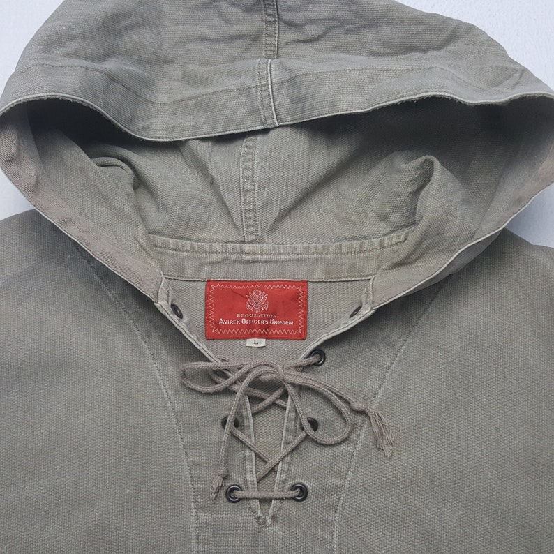Puede incluir: Una camisa con capucha de lona verde oliva con escote con cordones. La camisa tiene una etiqueta roja con texto blanco "REGULATION AVIREX OFFICER'S UNIFORM". La capucha est&aacute; unida al cuello.