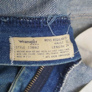 May include: Close-up of blue denim jeans with a visible label. The label says "Wrangler Brand" and includes details such as "Waist 33" and "Length 34". The jeans are made in the USA.