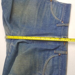 May include: Close-up of blue denim jeans with a measuring tape showing a width of approximately 35.5 cm. The jeans have classic pocket designs and metal button details. The denim has a faded, worn appearance.