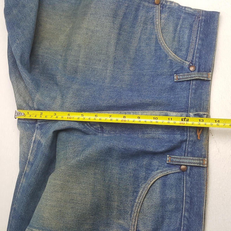 May include: Close-up of blue denim jeans with a measuring tape showing a width of approximately 35.5 cm. The jeans have classic pocket designs and metal button details. The denim has a faded, worn appearance.