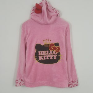 May include: Pink hooded sweatshirt featuring a Hello Kitty graphic on the back. The graphic includes a crown and bow. The hood has a red bow and a patterned lining. The cuffs have a pink and brown animal print.