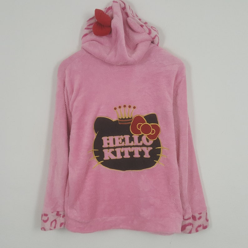 May include: Pink hooded sweatshirt featuring a Hello Kitty graphic on the back. The graphic includes a crown and bow. The hood has a red bow and a patterned lining. The cuffs have a pink and brown animal print.