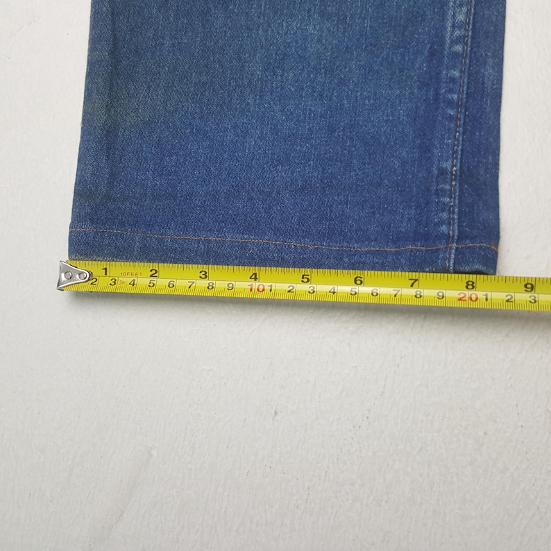 May include: Close-up of blue denim fabric with a measuring tape showing a length of approximately 20 cm. The tape measure is yellow with black markings, and the fabric has visible stitching.