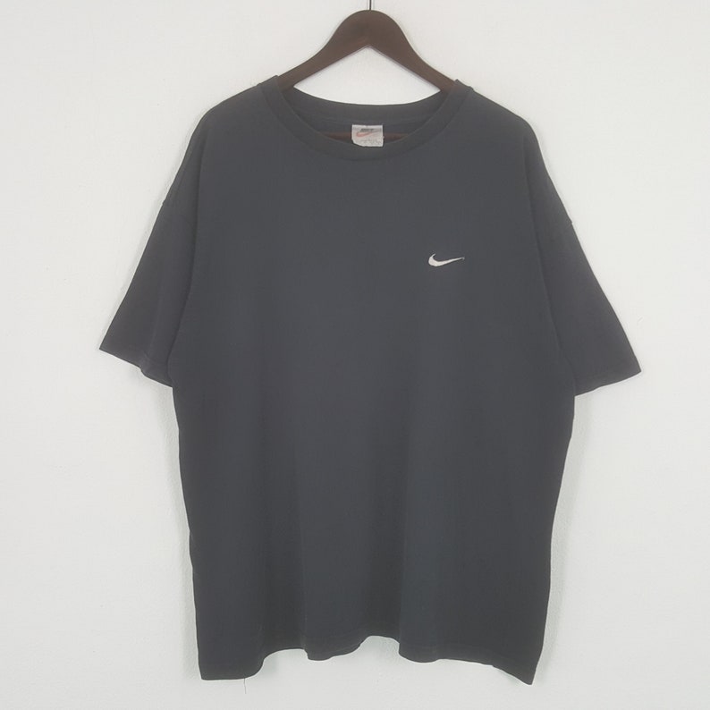 Peut inclure: Un t-shirt gris fonc&eacute; avec un petit logo Nike blanc sur la poitrine gauche. Le t-shirt a des manches courtes et un col rond. Le t-shirt est suspendu &agrave; un cintre en bois.