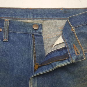 May include: Close-up of blue denim jeans with a partially unzipped brass zip and a button closure. The jeans have a classic five-pocket design and show signs of wear, suggesting a vintage style. The Wrangler brand is visible on the button.
