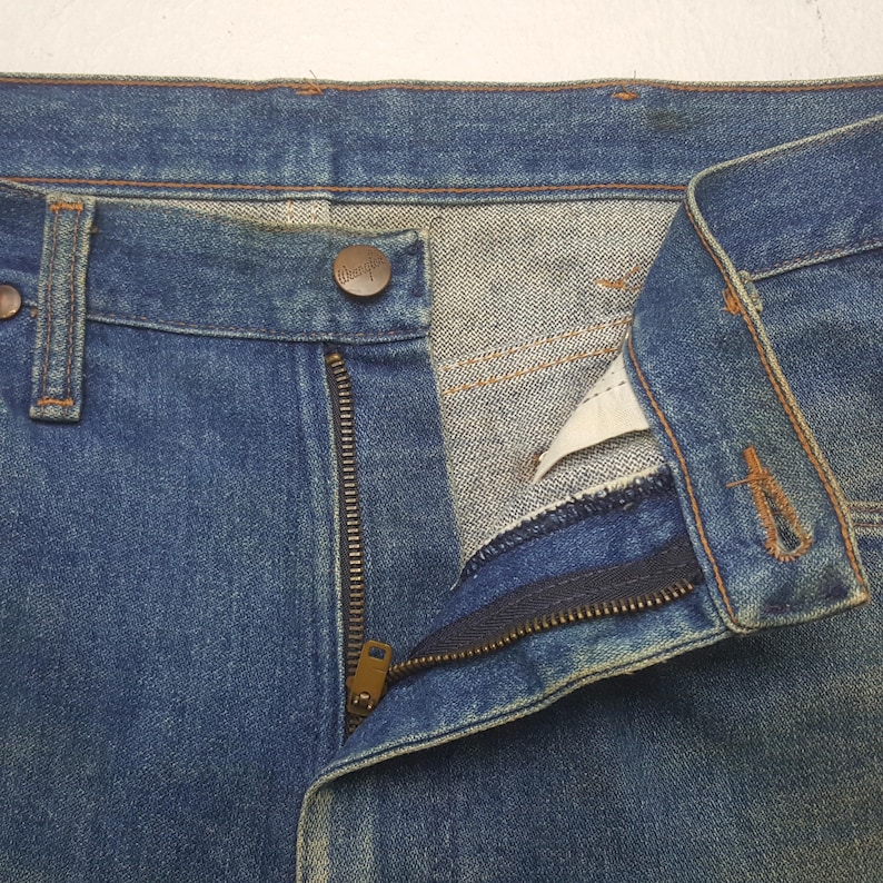 May include: Close-up of blue denim jeans with a partially unzipped brass zip and a button closure. The jeans have a classic five-pocket design and show signs of wear, suggesting a vintage style. The Wrangler brand is visible on the button.
